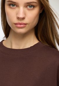 Brązowy sweter z prążkowanym dekoltem, o gładkiej fakturze. Modelka ma proste włosy i neutralny makijaż, skupiając się na odzieży.