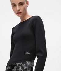 Pulover tricotat negru cu mâneci lungi și decolteu rotund. Prezintă un logo alb subtil pe partea de jos și este asociat cu o fustă/pantaloni cu un model.