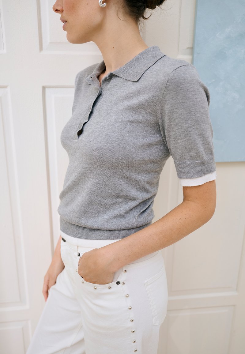 Vrouw draagt een grijs nauwsluitend poloshirt met korte mouwen en witte broek met metalen studs, staand met één hand in de zak tegen een witte paneelachtergrond.