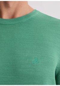 Grüner Strickpullover mit rundem Ausschnitt und gerippten Bündchen, mit einem kleinen geprägten Logo auf der Brust. Strukturierte Textur und figurbetontes Design.