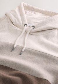 Svetlo bež pulover s kapuco s kremastim in bež blok dizajnom. Ima vrvico z metalnimi konicami in mehak, teksturiran material.