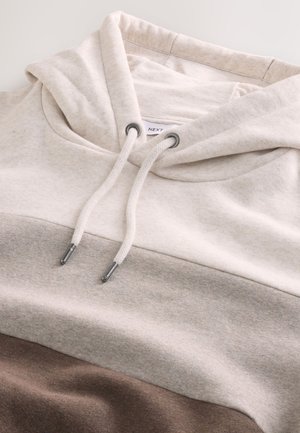 Svetlo bež pulover s kapuco s kremastim in bež blok dizajnom. Ima vrvico z metalnimi konicami in mehak, teksturiran material.