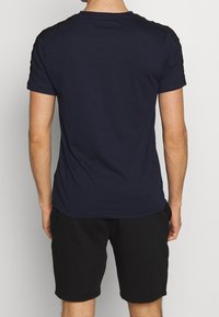 Navy blue short-sleeve t-shirt with black shoulder accents, made from smooth fabric. Worn with black shorts, showing a casual fit.