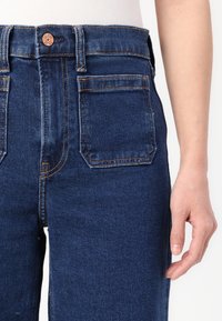 Tmavě modré džíny z denimu s vysokým pasem, pěti kapsami a měděným knoflíkem na pase. Tkanina má hladkou strukturu.