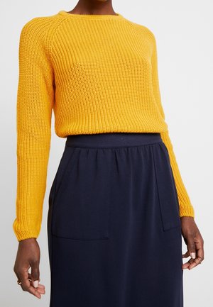 Pull en maille jaune à manches longues associé à une jupe taille haute bleu marine avec poches plaquées à l'avant.