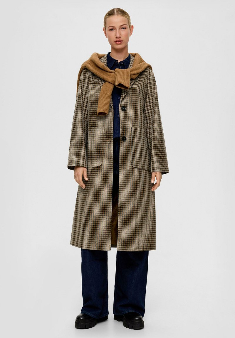 Bruine houndstooth jas met een relaxte pasvorm, grote knopen, voorzakken en verlaagde schouders. Een karamelskleurige trui rond de nek geknoopt.