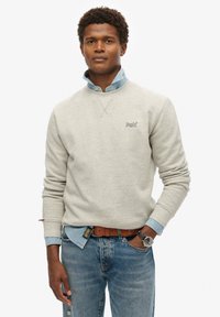 Lys grå sweatshirt med ribbet rund hals, der har et lille broderet logo. Laget over en lys denimskjorte, båret med blå jeans.
