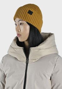 Beanie a coste di colore giallo senape con un'etichetta nera, indossato sopra una giacca a vento beige con un ampio cappuccio e chiusura con zip.