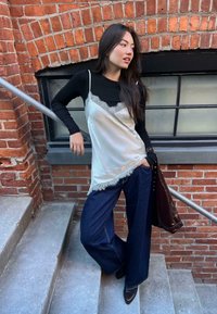 Žena v bielej tielku s čipkovým lemom, oblečenej nad čiernym dlhým rukávom, širokými džínsami a čiernymi čižmami, opierajúca sa o zábradlie na schodoch pri tehlovej stene.