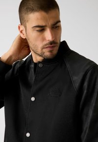 Zwarte jas met een gladde textuur, metalen drukknoopsluitingen en een reliëfmerk. Heeft een omslagkraag en contrasterende mouwaccenten.