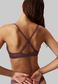 Reggiseno viola con schienale in pizzo e spalline incrociate regolabili, dotato di chiusura con gancio e motivo floreale sulla banda. Texture liscia, design aderente.