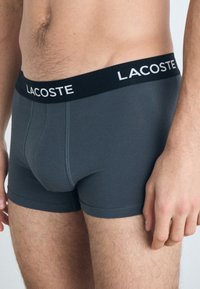 Graue Baumwollboxershorts mit einem schwarzen Bund, auf dem das "Lacoste"-Logo zu sehen ist. Das Design umfasst kontrastierende Nähte und eine enge Passform.