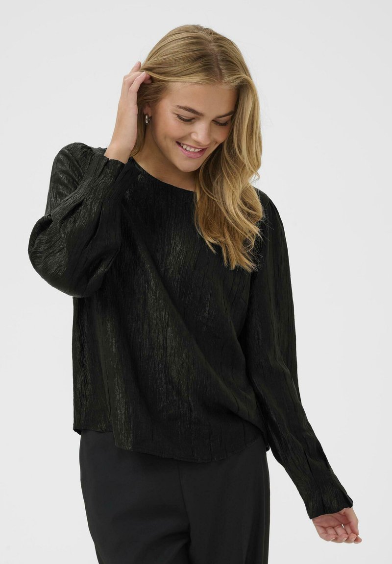 Giovane donna con lunghi capelli biondi che indossa una blusa nera a maniche lunghe con texture e pantaloni neri, sorridente e tocchiando i capelli.