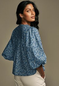 Bluzka z kwiatowym wzorem w odcieniach niebieskiego, z bufkami i okrągłym dekoltem. Miękka tkanina o fakturowanym wyglądzie, zestawiona z białymi spodniami.