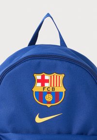 Sac à dos en tissu bleu avec un écusson FC Barcelone en évidence et un logo Nike jaune. Il a une forme arrondie avec une poignée supérieure et une fermeture zippée.