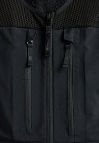 Czarna kamizelka z teksturowanym górnym materiałem siateczkowym i gładką dolną częścią, z dwoma wyraźnymi pionowymi zamkami błyskawicznymi oraz miękką podszewką.