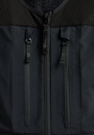 Schwarze Weste mit einem strukturierten Mesh-Oberteil und glattem Stoffunterteil, zwei auffälligen vertikalen Reißverschlüssen und einem weichen Innenfutter.