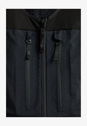Sort vest med et tekstureret mesh-overdel og glat stof i bunden, med to fremtrædende lodrette lynlåse og en blød inderside.