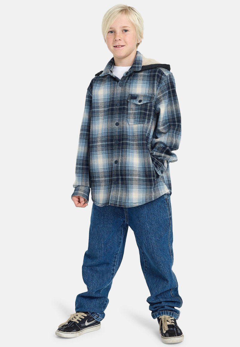 Camicia di flanella a quadri blu e bianchi con colletto e tasche laterali, abbinata a jeans blu larghi e sneakers nere.