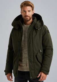 Tmavě zelená zateplená bunda s kapucí, zipem a klopy na druky, bočními kapsami a pod ní šedý svetr s texturou.