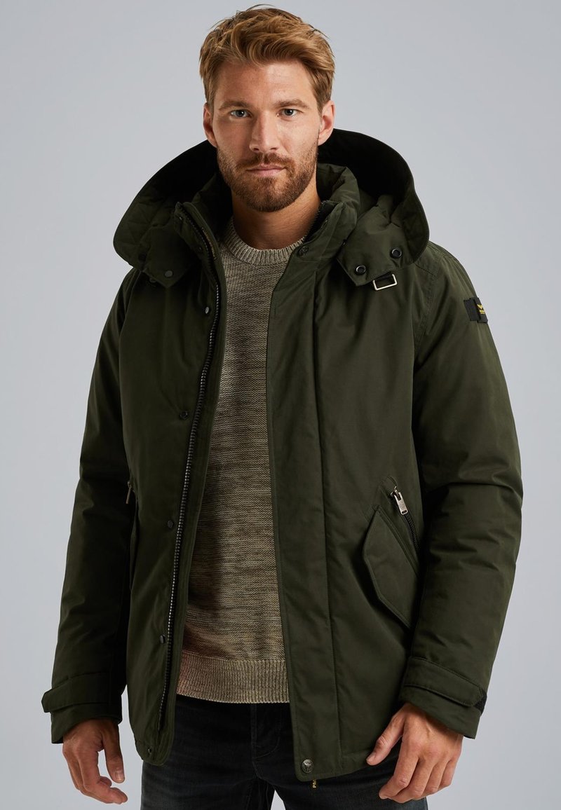 Tmavě zelená zateplená bunda s kapucí, zipem a klopy na druky, bočními kapsami a pod ní šedý svetr s texturou.