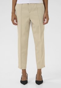 Pantalons beiges sur mesure à coupe droite, avec un devant plissé et des fentes latérales à l'ourlet, associés à des escarpins noirs à bout pointu.