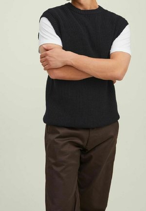 Czarny dzianinowy bezrękawnik na białej koszuli z krótkim rękawem, zestawiony z brązowymi spodniami. Tekstura jest prążkowana, a krój luźny.