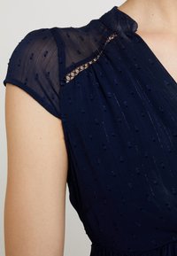 Šaty z průhledné tkaniny v námořnické modré barvě s tečkovanou texturou, s krátkým rukávem a malým krajkovým výřezem blízko ramenního švu.
