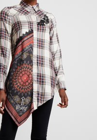Camisa abotonada con un patrón de cuadros en beige, burdeos y azul marino, con un dobladillo asimétrico. Los detalles incluyen bordado floral y un panel con un diseño.