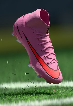 Crampon de football Nike rose avec logo swoosh rouge en suspension au-dessus d’un terrain en herbe, avec une ligne blanche visible en arrière-plan.