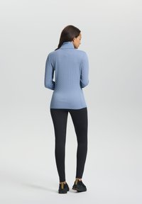 Hellblaues, strukturiertes Sportoberteil mit hohem Kragen, kombiniert mit schwarzen Leggings und dunklen Sneakers, von hinten betrachtet.