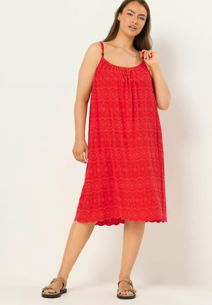 Mujer con vestido midi rojo sin mangas con ojales y dobladillo festoneado, y sandalias beige, de pie contra un fondo claro y liso.