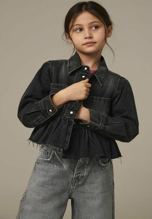 Chaqueta de mezclilla negra con cierre de botones, dobladillo acampanado y mangas largas. Combinada con pantalones de mezclilla gris desgastados. Diseño simple y casual.