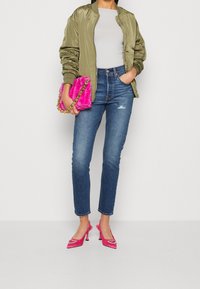 Chaqueta bomber verde oliva, blusa blanca ajustada, jeans azul clásicos, zapatos de tacón rosa y un bolso de piel rosa suave con cadena dorada.