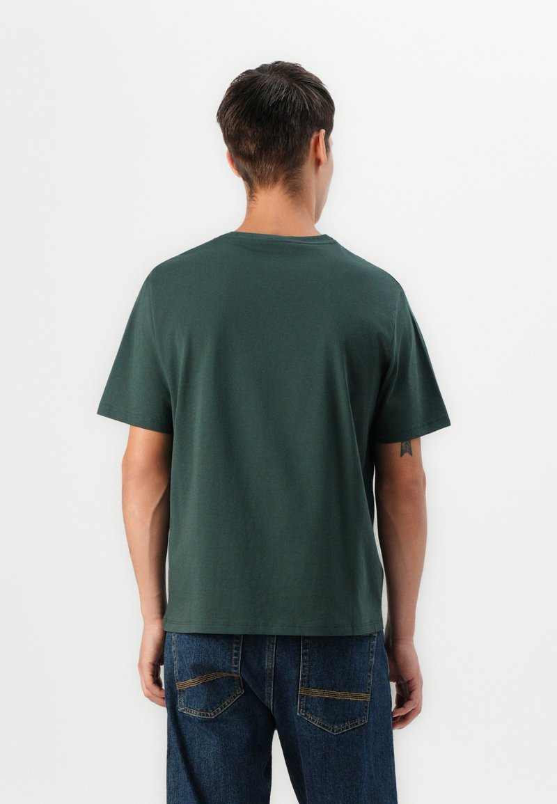 T-shirt in cotone verde scuro a maniche corte con collo rotondo, taglio dritto e texture liscia. Il modello lo indossa con jeans in denim blu.