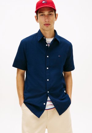 Ung mand iført en rød kasket, navy kortærmet button-up skjorte, stribet T-shirt og beige bukser, stående med hænderne i lommerne.