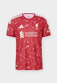 Κόκκινη φανέλα ποδοσφαίρου της Liverpool FC με λευκά αφηρημένα σχέδια, λογότυπο Adidas, χορηγό Standard Chartered και λευκές ρίγες στους ώμους και τα μανίκια.