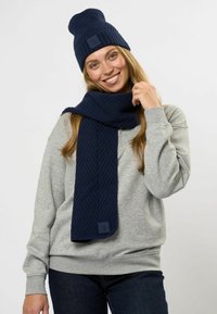 Mørkeblå strikket hue med ribbet tekstur og logo patch, parret med et matchende tørklæde med fiskebensmønster. Grå sweatshirt under.