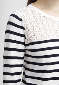 Pruhovaný svetr v námořnické modré a bílé barvě, s krajkovou yoke s kruhovými vzory, dlouhými rukávy a kulatým výstřihem. Měkká textury.