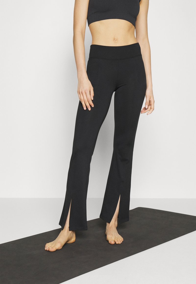 Femme debout pieds nus sur un tapis de yoga noir, portant des leggings évasés courts noirs avec des fentes à l'avant et un soutien-gorge de sport noir.