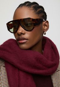 Occhiali da sole extralarge con montatura in tartaruga marrone scuro e lenti verde scuro. Una morbida sciarpa color maroon si appoggia attorno al collo.