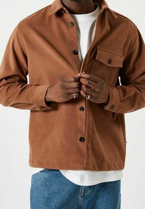 Chemise en polaire marron avec des boutons noirs, poche poitrine et coupe décontractée, portée par-dessus un t-shirt blanc et un jean bleu.