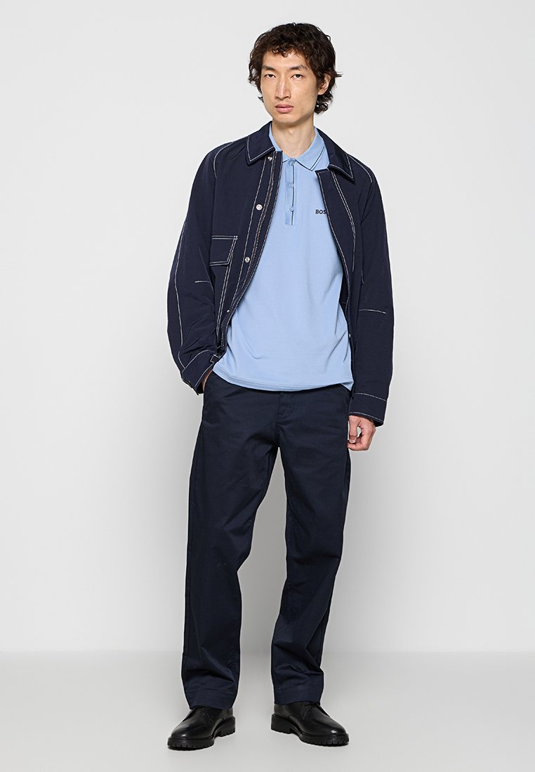 Junger Mann in einer dunkelblauen Jacke mit weißer Naht, hellblauem Poloshirt, dunkler Hose und schwarzen Schuhen, der vor einem einfarbigen grauen Hintergrund steht.