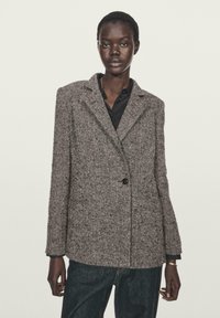 Blazer grigio a spina di pesce con chiusura a un bottone, rever a punta e due tasche frontali. Indossato sopra una camicia nera, abbinato a jeans scuri.