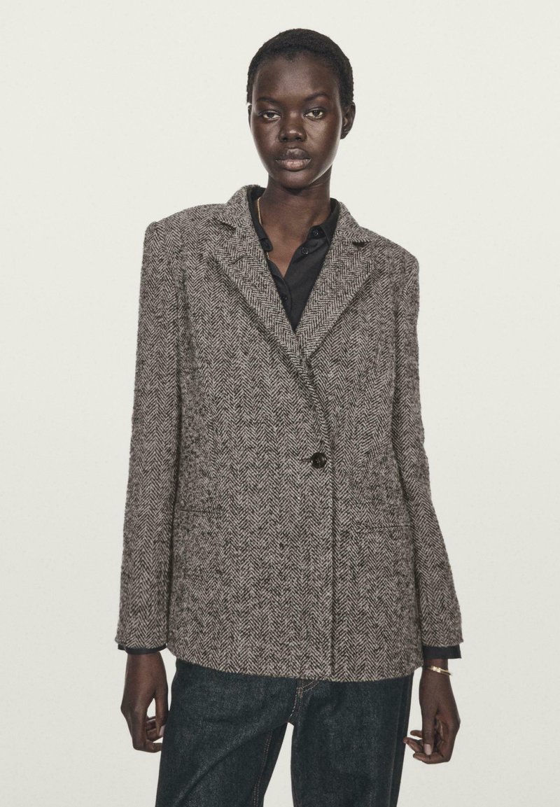 Blazer grigio a spina di pesce con chiusura a un bottone, rever a punta e due tasche frontali. Indossato sopra una camicia nera, abbinato a jeans scuri.