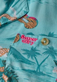 Koszula z krótkim rękawem w jasnoturkusowym materiale z tropikalnym wzorem, przedstawiającym palmy i gitary. Różowe haftowane logo i kremowe guziki.