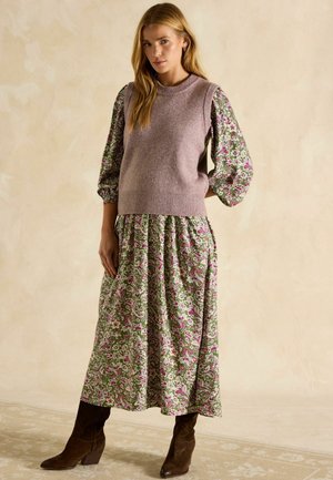 Gestricktes, lavendelfarbenes, ärmelloses Oberteil über einem floralen, grünen Maxikleid mit rosa Akzenten. Das Modell trägt dazu braune Wildleder-Stiefeletten.