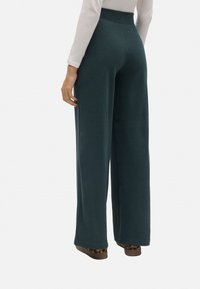 Pantalones anchos de color teal oscuro en una tela suave con una cintura alta. Presenta un dobladillo terminado con un borde limpio, combinados con zapatos de estampado de leopardo marrón.