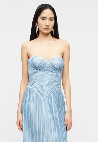 Γυναίκα με μακριά σκουρόχρωμα μαλλιά, που φοράει αμάνικο, ριπέν γαλάζιο φόρεμα και μακριά κρεμαστά σκουλαρίκια από κρύσταλλο, πάνω σε απλό φόντο.