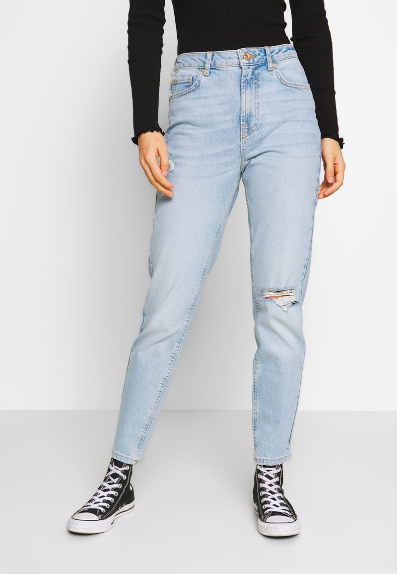 Personne portant un jean bleu clair déchiré, des baskets montantes noires et un haut à manches longues noir, debout devant un fond uni.